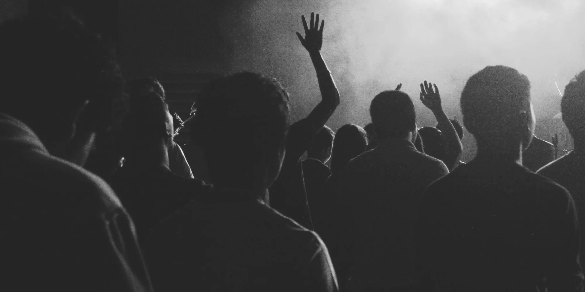 A tightly packed concert room seen from behind the crowd, with hands raised into the stage haze.